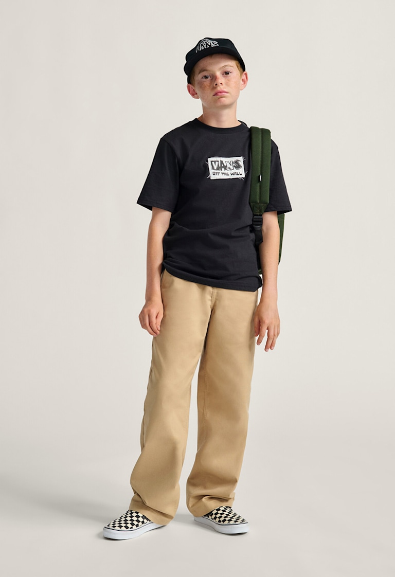 A boy poses wearing a Vans black graphic t-shirt, brown chinos, a green backpack, and white and black Checkerboard Classic Slip-On.