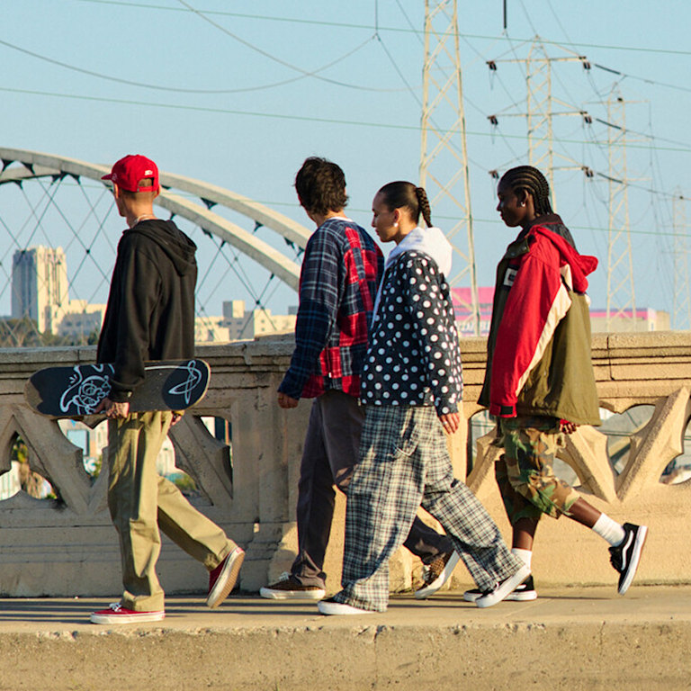 A group of friends cross a bridge wearing Vans shoes mixed in with thrifted and vintage styles during golden hour.