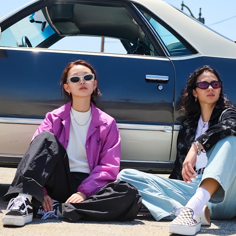 Two women sit on the asphalt next to a vintage car wearing Vans clothes, Old Skool, and Checkerboard Classic Slip-On.
