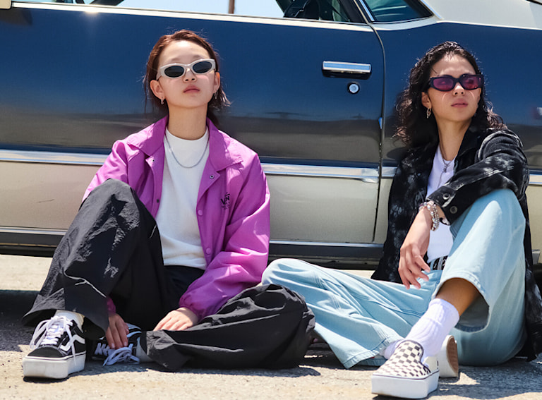 Two women sit on the asphalt next to a vintage car wearing Vans clothes, Old Skool, and Checkerboard Classic Slip-On.