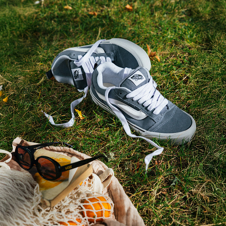 A pair of gray and white Knu Skool lie in the grass with their laces untied next to a grocery bag for a picnic.