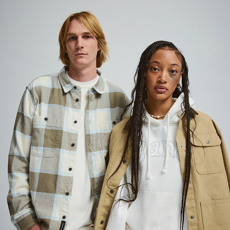 A young man in a Larkspur Plaid Flannel Shirt stands next to a young woman in a tan Drill Chore Coat in a photography studio.