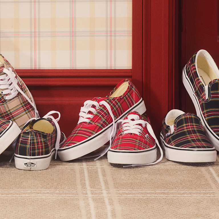 Several pairs of plaid Premium Authentic and Premium Classic Slip-On lie on the carpeted floor of a sitcom set.