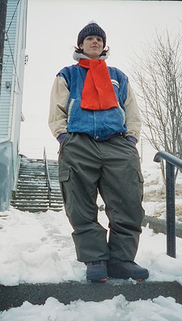 Vans athlete and professional snowboarder Jill Perkins standing in the snow in gray beanie, red scarf, denim jacket, and gray snow pants.