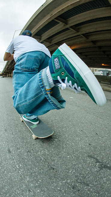 A close-up of green and blue Half Cab shoes as a skateboarder pushes off the ground on his skateboard.