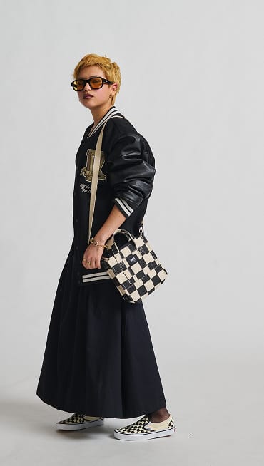 A woman posing and wearing a black and white Checkerboard bag with a Vans varsity-style jacket and a black dress.