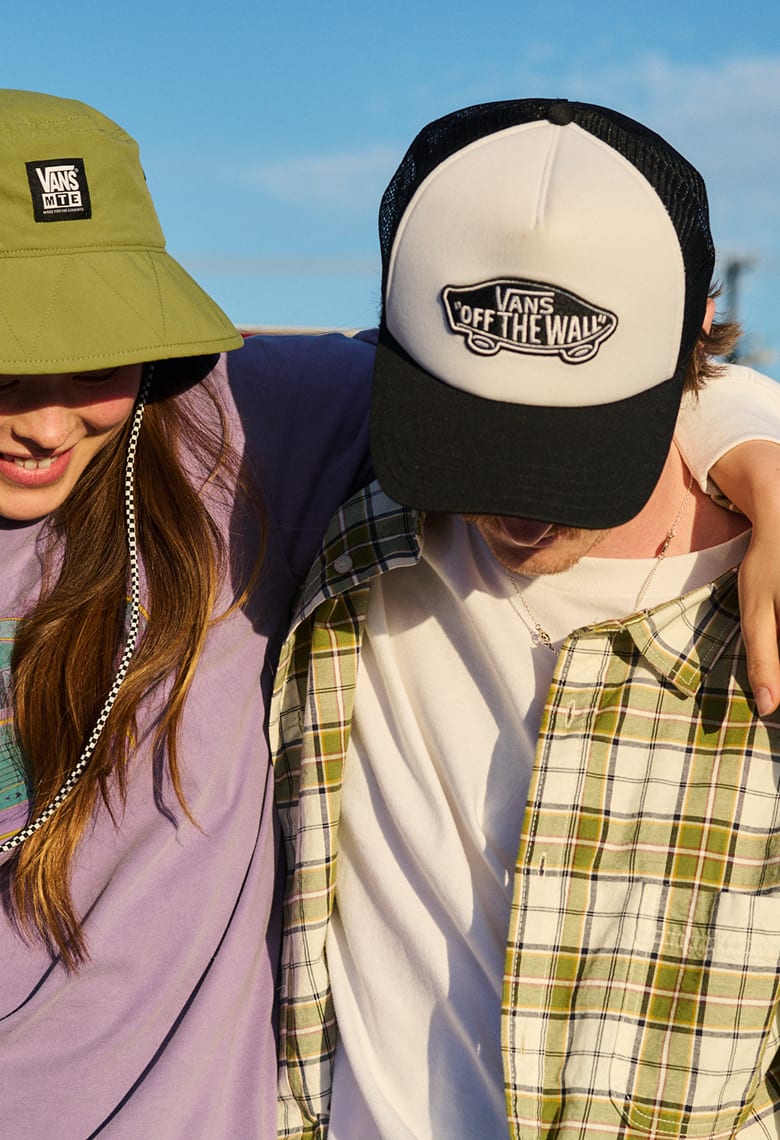 Two people wearing Vans hats with their arm around each other's shoulders while looking down. 