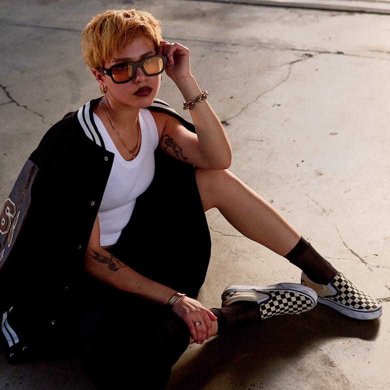 A woman with short bleached hair sitting on concrete wearing a Vans varsity-style jacket and black and white Checkerboard Slip-On.