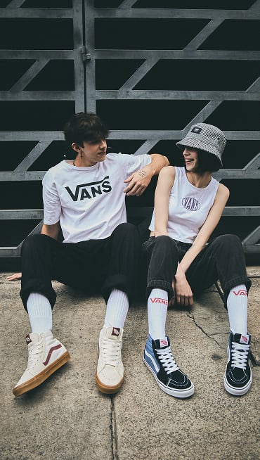 A boy and a girl sitting on the ground against a gate wearing clothes with the Vans logo and Sk8-Hi shoes.