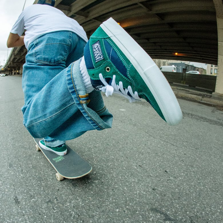 A close-up of green and blue Half Cab shoes as a skateboarder pushes off the ground on his skateboard.