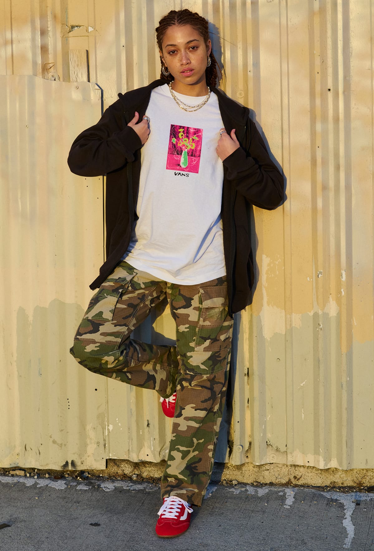 A woman leaning on a shipping crate and wearing red Super Lowpro with camo pants and a black hoodie.