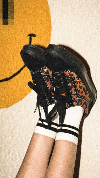 A person resting their feet upwards on a wall wearing the black and leopard print Premium Old Skool from the Premium Old Skool Music collection with a big smiley-face poster behind them.