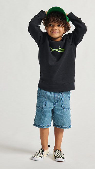 A child in a black sweatshirt with a green graphic, denim shorts, and Kids Classic Slip-On shoes in Checkerboard pattern.