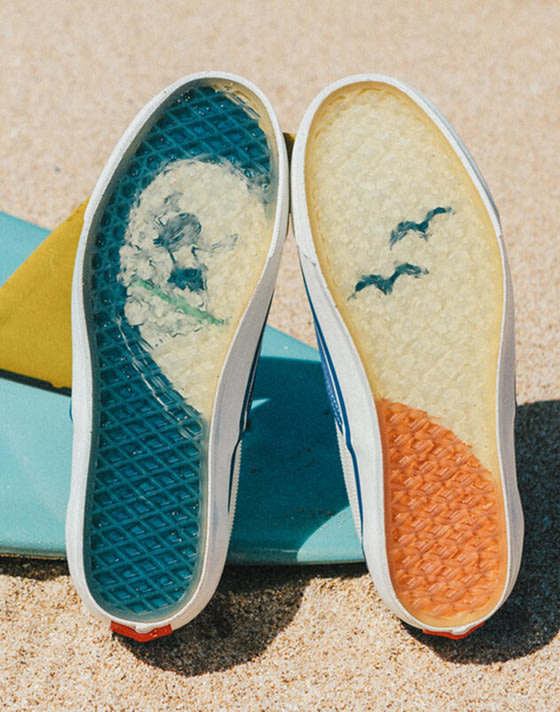 A pair of Authentic Reissue sneakers in Thalia Blue stand upright, soles facing up in focus, against a multicolored surfboard lying in the sand. The soles feature a surfing motif with a wave, surfer, and seagulls.
