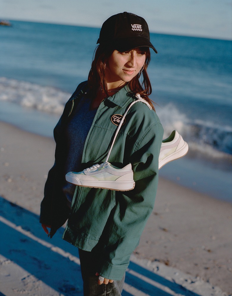 A woman looks down the beach in a green Vans jacket and a pair of UltraRange 2.0 slung over her shoulder by the laces.