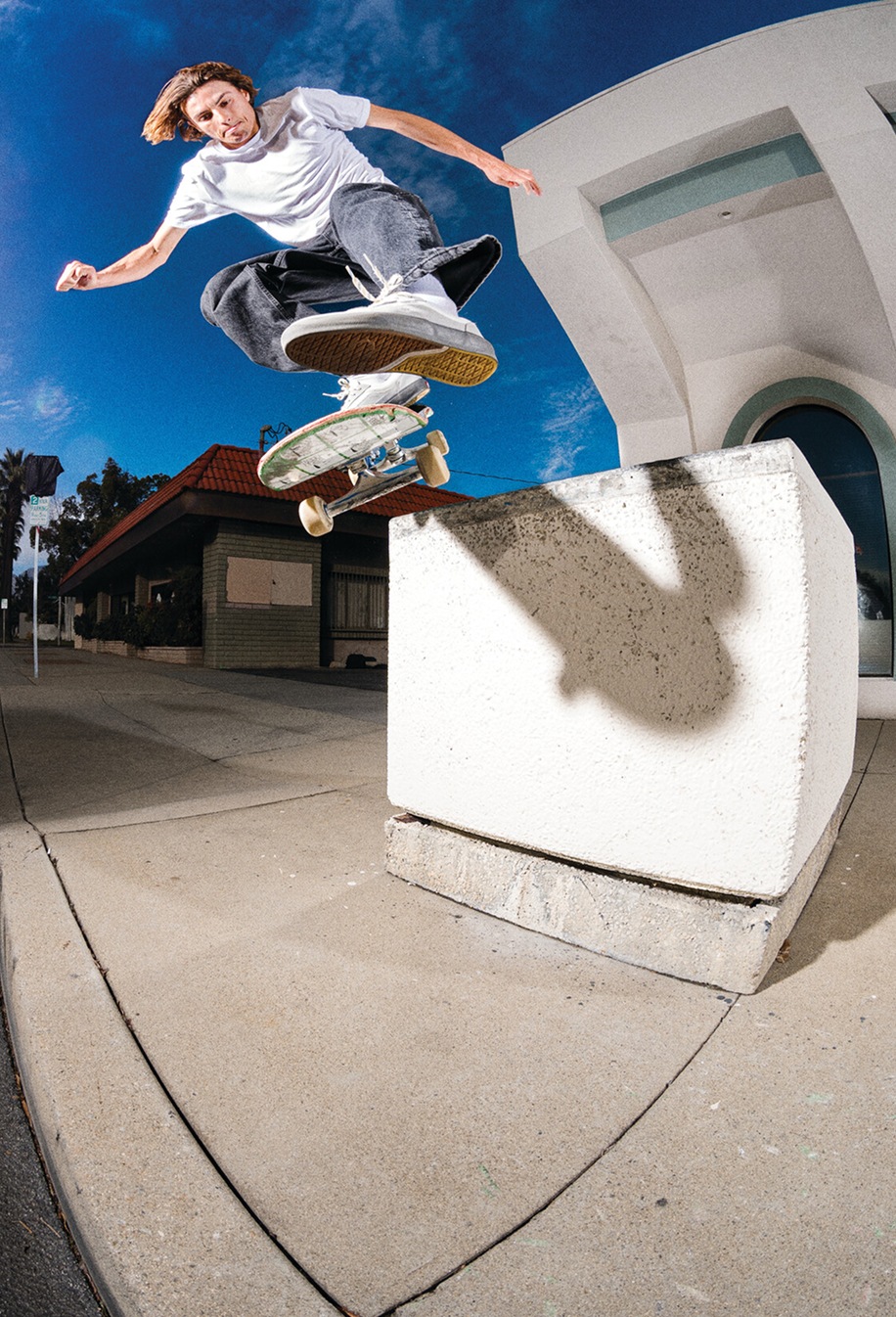 A well-lit shot of Curren jumping to grind on a concrete block wearing a white t-shirt, black jeans, and white Curren shoes.