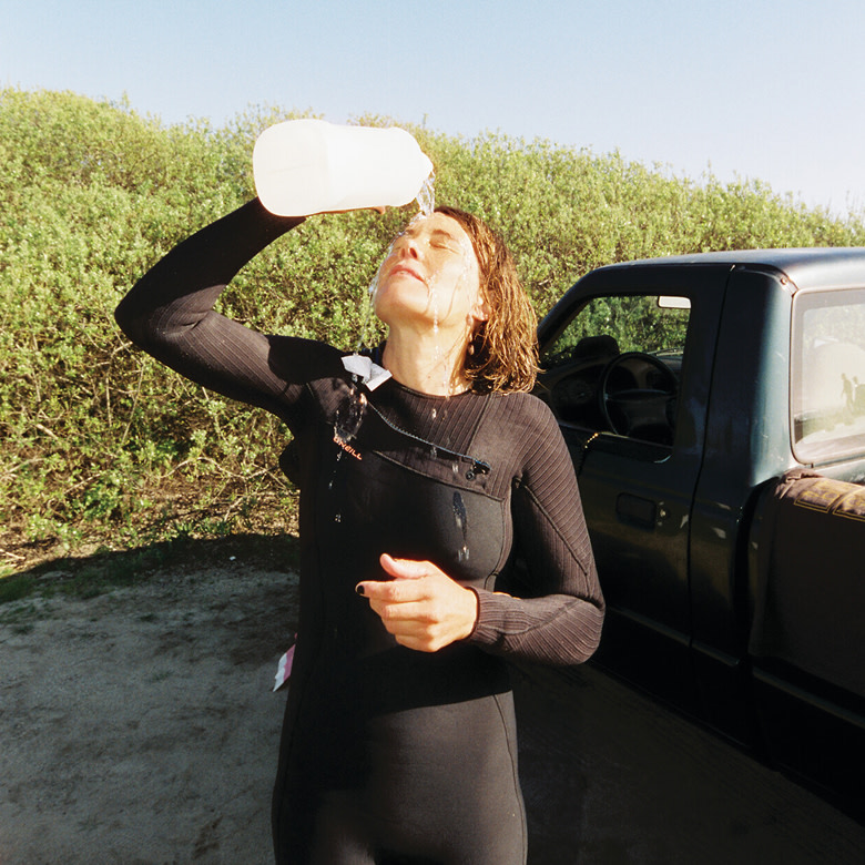 A surfer wearing a wetsuit pours water on their face near the beach.