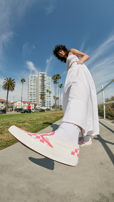 A woman throws her foot towards the camera wearing Honeysuckle Pink Checkerboard Classic Slip-On paired with white socks near the beach.