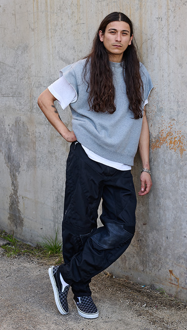 A man leans on a cracked concrete wall wearing dark jeans and gray and black Checkerboard Classic Slip-On.