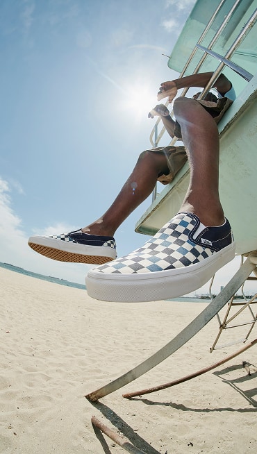 A close-up of someone sitting on a lifeguard tower wearing navy blue and white Checkerboard Classic Slip-On with the beach and sky in the background.