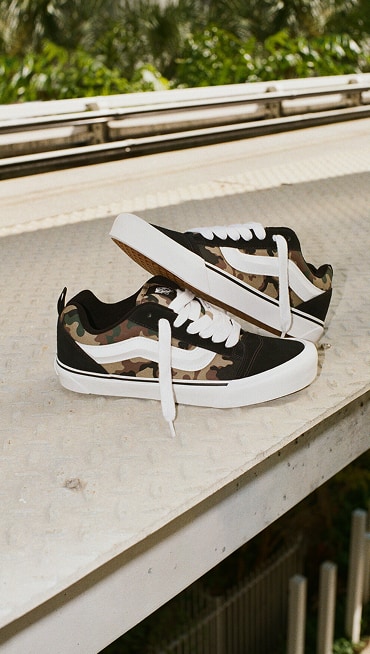A pair of camo Knu Skool lie on a metal floor stacked on top of each other to create a gritty urban vibe.