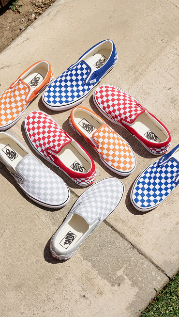A variety of Checkerboard Classic Slip-On lie on the concrete in various colors.