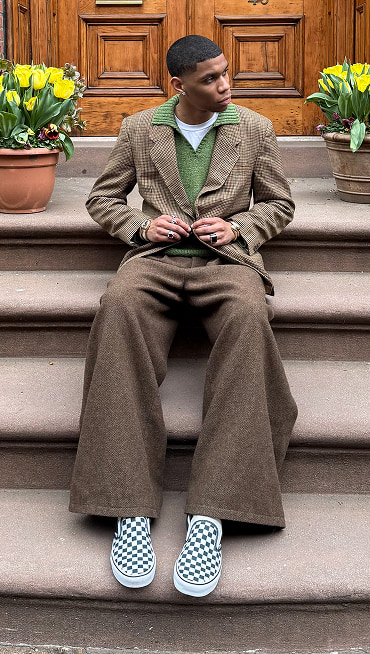 @nassnyc sits on a staircase wearing a brown houndstooth blazer, baggy tweed pants, and white and black Checkerboard Classic Slip-On.