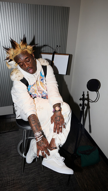 Lil Uzi Vert wears white pants and a white hoodie, showing a silver anklet over his white Old Skool shoes in his studio.