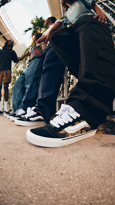 A close-up of a pair of camo Knu Skool on a man wearing black jeans with a group of young people in the background under a tram line.