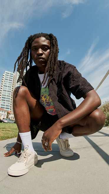 A ground-up shot of a man wearing baggy blue jeans and Skate Old Skool Shoes with his foot on a skateboard in a park.