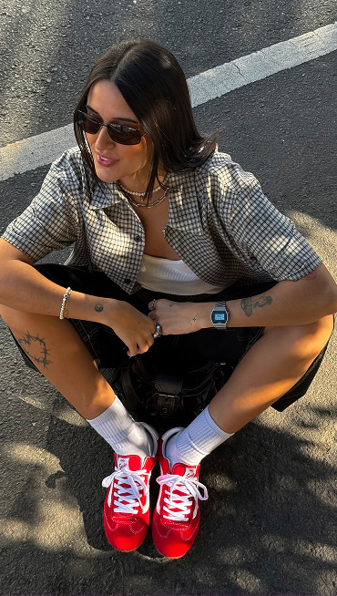 A woman poses sitting on the street, wearing sunglasses, a checkered button-down, black shorts, and racing red Vans Super Lowpro shoes.