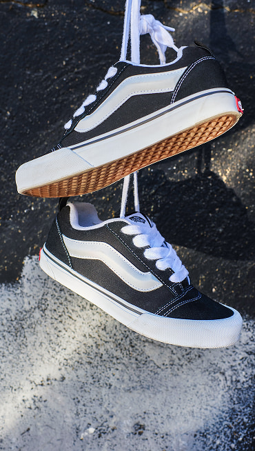 A pair of black and white Vans Knu Skool shoes hang by the laces against a half-painted wall.