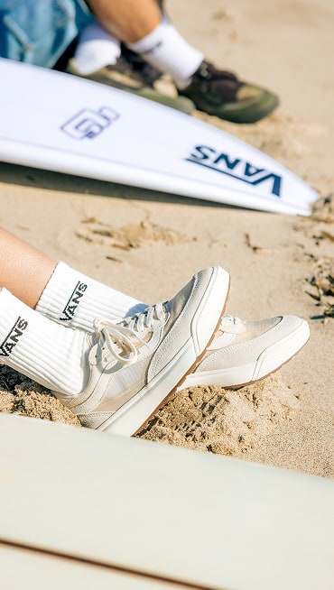 A woman wears a pair of UltraRange 2.0 with her feet in the sand next two some surfboards.