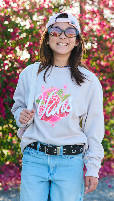 A young girl smiles for the camera with her sepia rose pink Old Skool Classic Backpack and Vans gray pullover in front of a rose bush.
