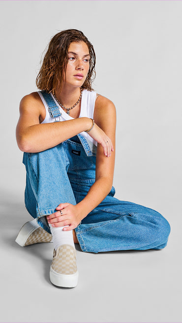 A young woman sits in a photography studio wearing Drew Rib Tank Top, denim blue Sirelle Overalls, and Checkerboard Platform Slip-On.