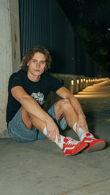 A man sits on the concrete and looks into the camera wearing a black t-shirt, denim blue jeans, and Racing Red Super Lowpro.