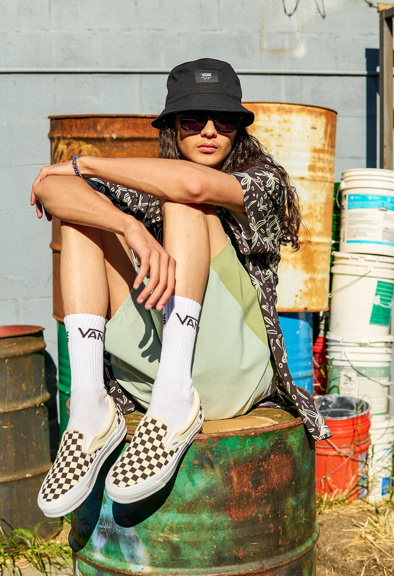 A young man sits on a rusty oil drum wearing black and white Checkerboard Classic Slip-On with white Vans crew socks and a black Vans bucket hat.