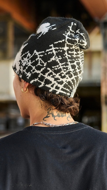 Close-up of the back of a man’s head with a neck tattoo wearing a Vans beanie with a spiderweb pattern.