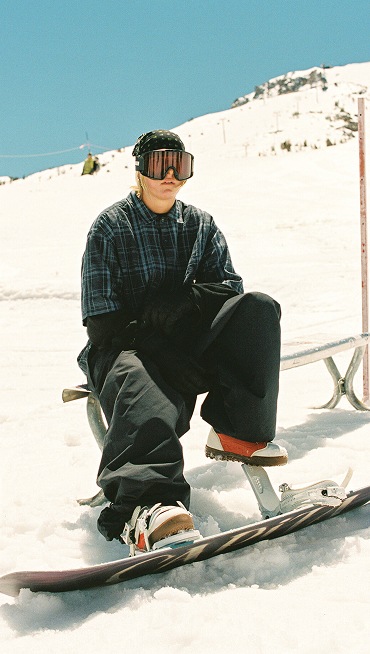Vans Snowboard Team Rider Mia Brookes wearing red and white Encore Pro snowboard boots and Vans clothes sitting on a metal bench on a ski slope.