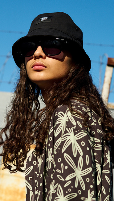 A man with long hair looks at the camera wearing a black and mint Hawaiian shirt and a black Vans bucket hat.