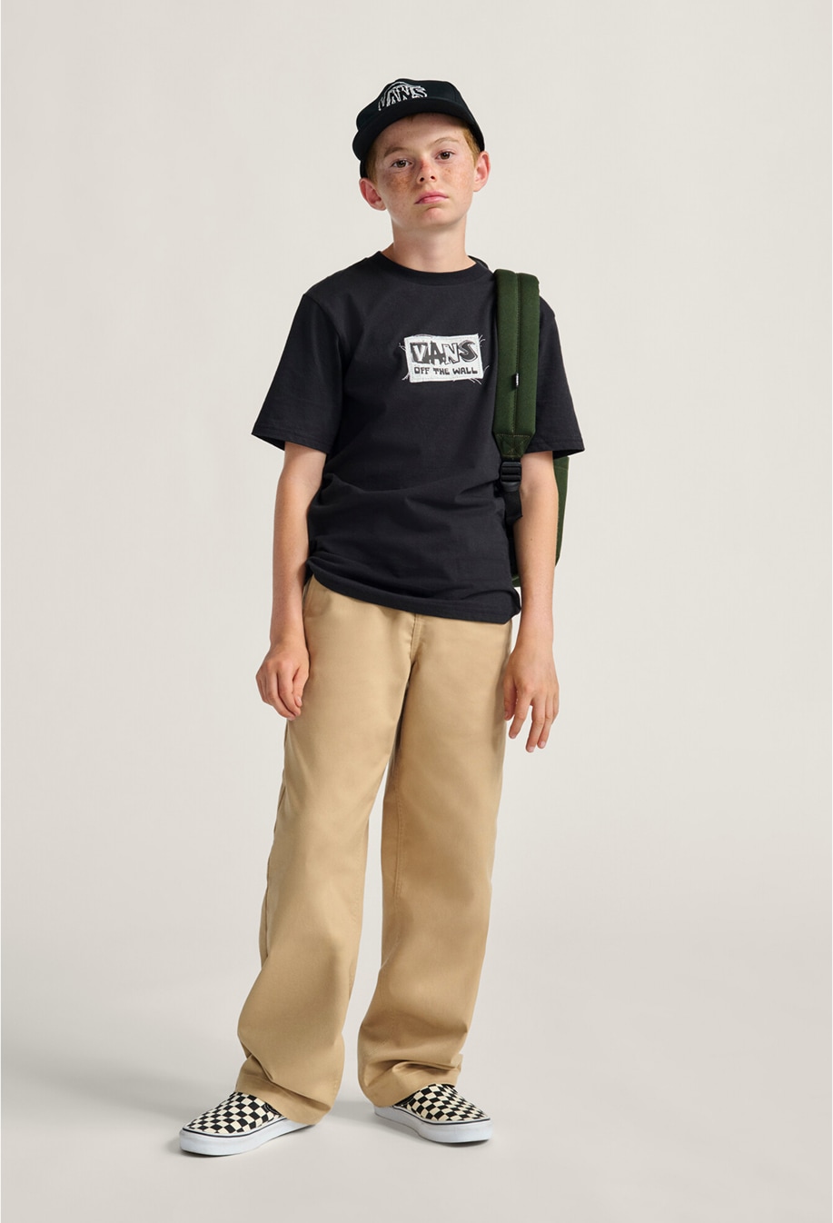 A boy poses wearing a Vans black graphic t-shirt, brown chinos, a green backpack, and white and black Checkerboard Classic Slip-On.