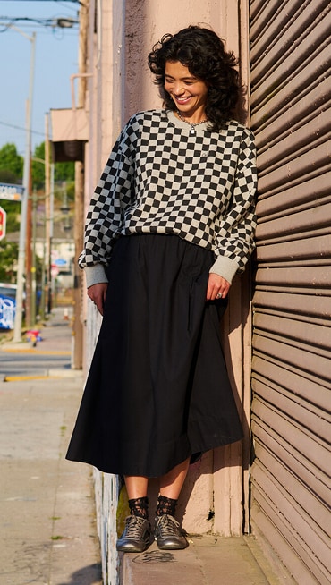 A young woman wearing a black Francesca Full Skirt and the Checkerboard Retro Crew Sweatshirt looks down at her black and silver Super Lowpro.