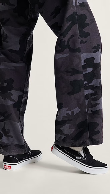 A knee-down shot of someone testing the fit of white and black Classic Slip-On paired with black and gray camo pants.