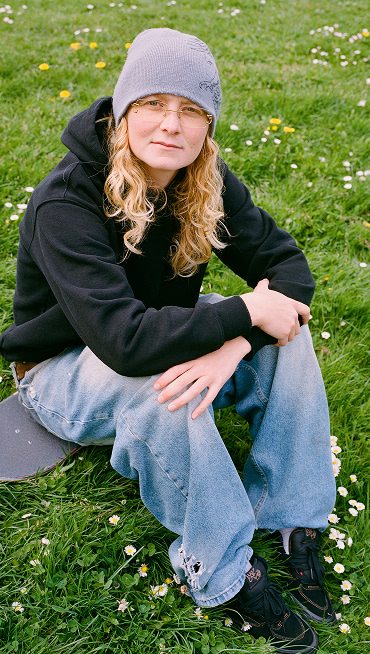 Vans Skate Team Rider Una Farrar looks into the camera and sits on a skateboard in the grass wearing black Skate Half Cab.