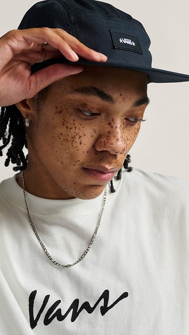 A close-up of a man in a black Vans five-panel hat and white t-shirt in a photography studio.