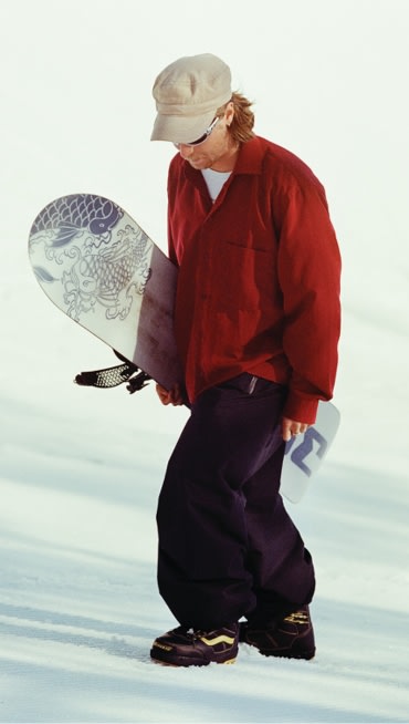 Rene Rinnekangas wearing Vans Invado Pro Snowboard boots, a red jacket, black pants, and a beige cap walks on snow while holding a snowboard.
