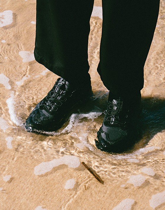 A knee-down shot of someone wearing the black Crosspath XC GORE-TEX and standing in a shallow wave on a sunny beach.