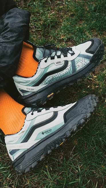 A close-up of someone wearing blue Crosspath XC GORE-TEX with black pants and orange socks on the grass.