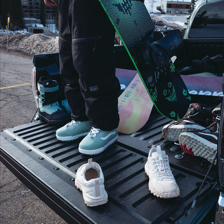 A young woman stands in the bed of a truck wearing all black snowboard gear and the Luna Pro with more boards and boots in the truck.