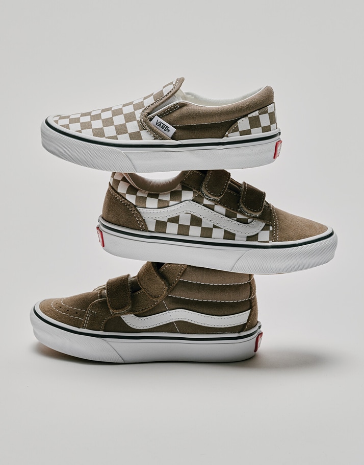 A brown Kids’ Checkerboard Classic Slip-On, brown Kids’ Old Skool, and brown Kids’ Sk8-Hi are stacked on each other in a photography studio.
